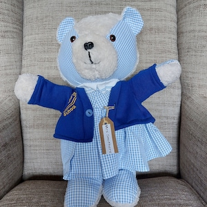 May include: A white teddy bear wearing a blue and white checkered dress, a blue jacket with a gold school logo, and a brown tag that says "Mrs. Smithers".