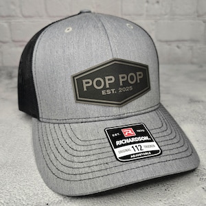 May include: A gray and black trucker hat with a black leather patch that says "Pop Pop Est. 2025". The hat is adjustable and has a Richardson 112 label.