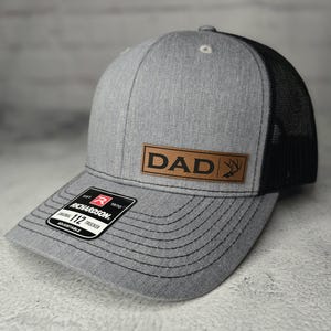 May include: A gray and black trucker hat with a brown leather patch that says "DAD" and a deer antler graphic. The hat is adjustable.