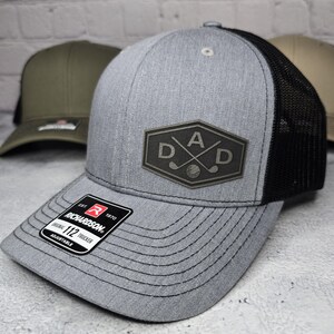 May include: A gray and black trucker hat with a black mesh back. The front of the hat features a black leather patch with the word "DAD" and crossed golf clubs.