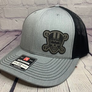 May include: Gray trucker hat with a black mesh back and a skull patch wearing a helmet with crossed wrenches. The hat has a curved brim and an adjustable strap. The patch features a skull wearing a helmet with stripes and crossed wrenches.