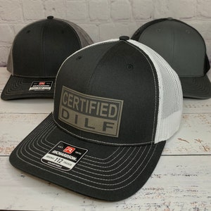 May include: A black and white trucker hat with a white mesh back. The hat has a brown patch on the front that says "CERTIFIED DILF".
