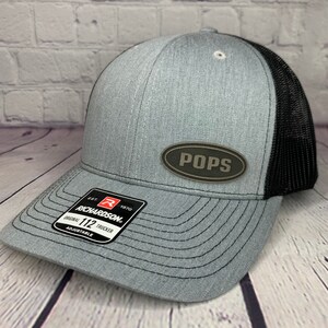 May include: A gray and black trucker hat with a brown oval patch that says "POPS" on it. The hat is made by Richardson 112 and is adjustable.