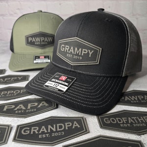 May include: Two black and green trucker hats with embroidered patches. The patches are in a hexagon shape and say "Pawpaw Est. 2025" and "Grampy Est. 2019". The hats are surrounded by several other patches with different names and dates.