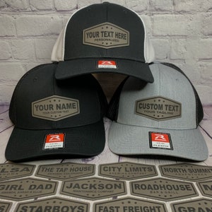 May include: Three different trucker hats with custom patches. The patches are brown with white text. The hats are black, gray, and white. The patches say "Your Text Here Personalized", "Your Name Your Custom Text", and "Custom Text Your Tagline".
