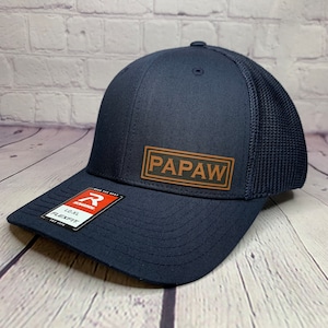 May include: A navy blue trucker hat with a brown leather patch that says "PAPAW" on it. The hat has a mesh back and a flexfit.
