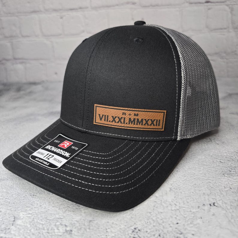 May include: Black and gray trucker hat with a brown leather patch that reads "R+M VII.XXI.MMXXII".