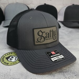 May include: A gray trucker hat with a black mesh back and a curved brim. The hat features a rectangular patch with the word "Salty" and "Matthew 5:13" printed on it. The hat is adjustable.