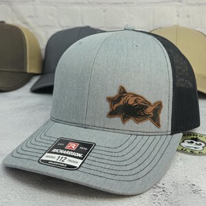 May include: Gray trucker hat with a black mesh back and a brown leather patch featuring a fish design. The hat has a curved brim and the text "RICHARDSON" on a tag.