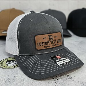 May include: A gray trucker hat with a white mesh back and a brown leather patch. The patch features the text "CUSTOM TEXT HERE" and a bull graphic. The hat is adjustable and has a Richardson tag.