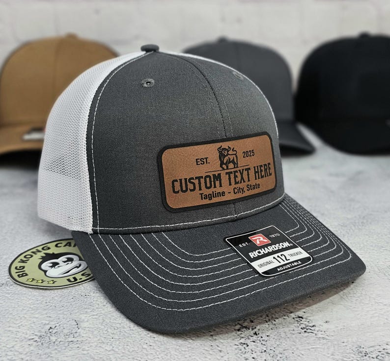 May include: A gray trucker hat with a white mesh back and a brown leather patch. The patch features the text "CUSTOM TEXT HERE" and a bull graphic. The hat is adjustable and has a Richardson tag.