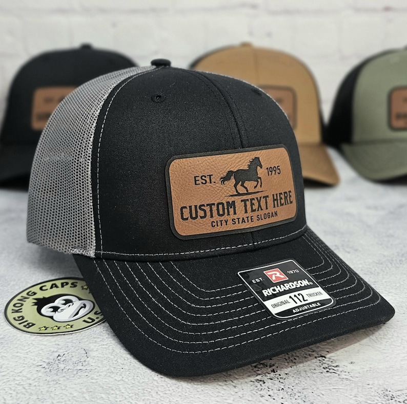K&ouml;nnte beinhalten: Schwarze Trucker-Cap mit grauem Mesh-R&uuml;cken und einem braunen Lederpatch. Der Patch zeigt eine Pferdesilhouette, den Text "EST. 1995" und Platz f&uuml;r individuellen Text. Die Cap hat einen gebogenen Schirm und wei&szlig;e N&auml;hte.