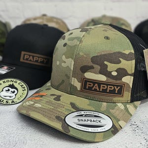 May include: A camouflage print snapback hat with a black mesh back and a brown leather patch that reads "PAPPY". The hat has a curved brim and a white tag that says "The Authentic Snapback". Other hats are visible in the background.