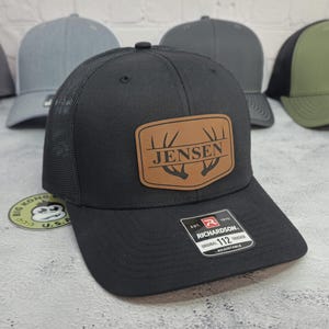 May include: Black Richardson 112 trucker hat with a brown leather patch that reads "Jensen" above a deer antler design. The hat has a mesh back and an adjustable strap. Other hats are visible in the background.