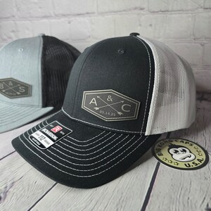 May include: Two black and white trucker hats with a black patch on the front. The patch has a hexagon shape with two arrows pointing towards each other and the letters A & C with the date 05.14.25 below. The hat on the right has a white mesh back and the hat on the left has a gray mesh back.