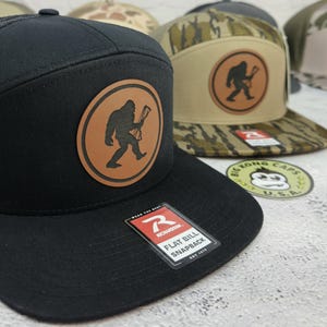 May include: Black flat bill snapback hat with a brown leather patch featuring a Bigfoot silhouette holding a bow and arrow. The hat has a Richardson tag. Other hats with camouflage and Bigfoot patches are visible in the background.