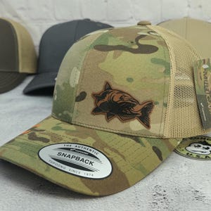 May include: Camouflage pattern snapback hat with a fish emblem. The hat features a brown leather patch with a fish design, a tan mesh back, and a curved brim. The hat has a snap closure and a white and black tag that says "SNAPBACK".