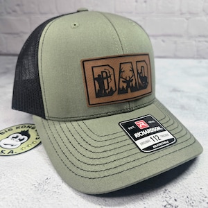 May include: Olive green trucker hat with a black mesh back and a brown leather patch that reads "DAD" with hunting-themed graphics. The hat has a curved brim and an adjustable strap.