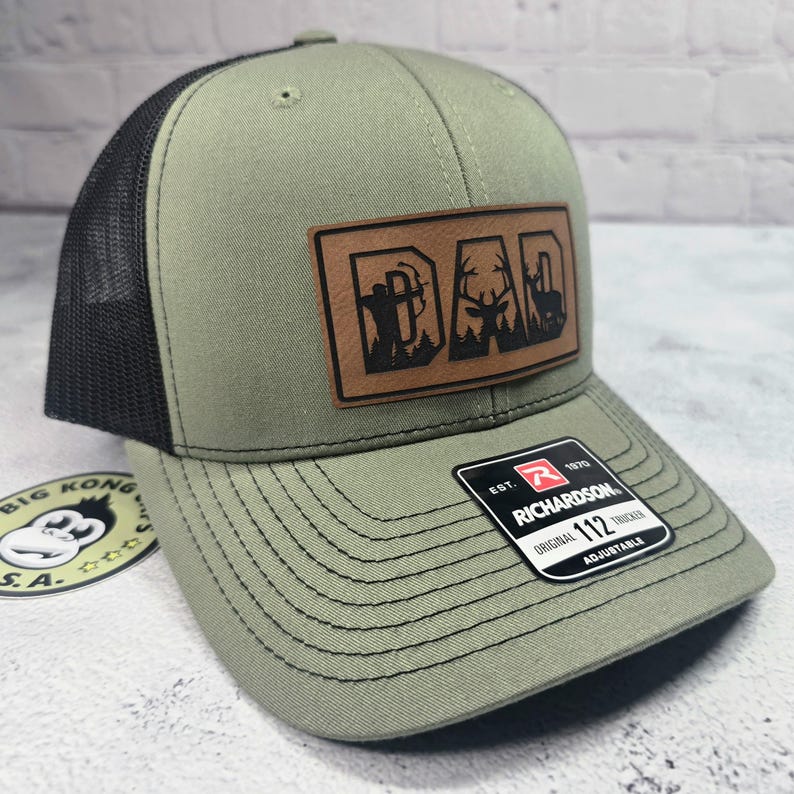 May include: Olive green trucker hat with a black mesh back and a brown leather patch that reads "DAD" with hunting-themed graphics. The hat has a curved brim and an adjustable strap.