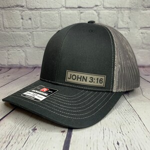 May include: A black and gray trucker hat with a gray mesh back. The hat has a black patch on the front that says "JOHN 3:16".