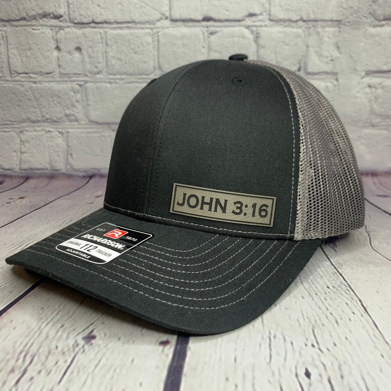 May include: A black and gray trucker hat with a gray mesh back. The hat has a black patch on the front that says "JOHN 3:16".