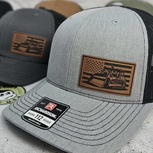 May include: Gray trucker hat with a black mesh back and a brown leather patch featuring an American flag and a bulldozer. The hat has a curved brim and an adjustable strap. The Richardson brand label is visible.