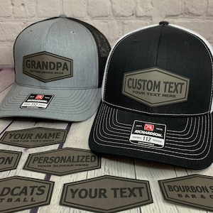 May include: Two black and gray trucker hats with a brown leather patch on the front. The patch on the left hat says "Grandpa Established 2024". The patch on the right hat says "Custom Text Your Text Here". There are several other brown leather patches with different text on them.
