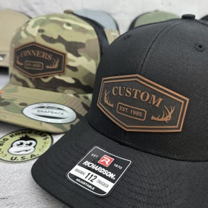 May include: Black and camouflage trucker hats with custom leather patches. One hat features "CUSTOM EST. 1995" and the other reads "CONNERS EST. 2025". Both hats have a Richardson 112 Trucker tag.