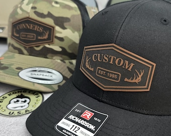 May include: Black and camouflage trucker hats with custom leather patches. One hat features "CUSTOM EST. 1995" and the other reads "CONNERS EST. 2025". Both hats have a Richardson 112 Trucker tag.