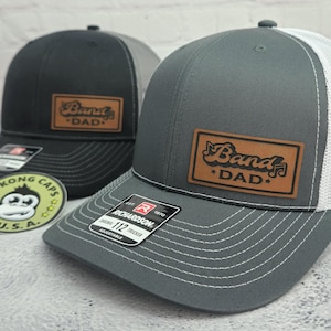 May include: Two trucker hats with a leather patch that reads "Band Dad". One hat is charcoal gray with a white mesh back, the other is black with a gray mesh back. Both have a curved brim and adjustable fit.