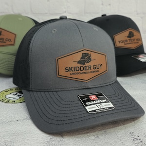 May include: Gray trucker hat with a black mesh back and a brown leather patch that reads "SKIDDER GUY LANDSCAPING & REMOVAL". The hat has a curved brim and an adjustable strap. A Richardson 112 Trucker label is visible.
