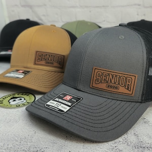 May include: A collection of trucker hats in various colors, including black, gray, and tan. Each hat features a leather patch with the word "SENIOR" and the year "2026". The hats have a curved brim and adjustable strap.