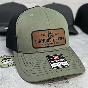 May include: Olive green trucker hat with a black mesh back. The hat features a brown leather patch with the text "DIAMOND X RANCH Chesterfield, Maine" and a bull graphic. The hat also has a Richardson 112 Trucker tag.