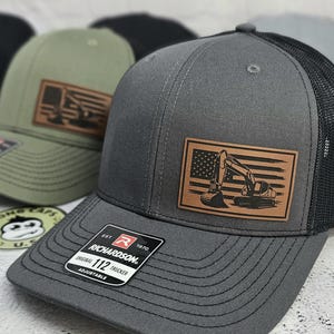 May include: Gray trucker hat with a black mesh back and a brown leather patch featuring an excavator over an American flag design. The hat has a curved brim and an adjustable strap. The Richardson brand label is visible.