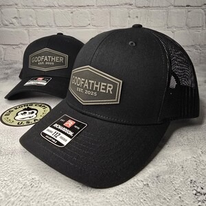 Puede incluir: Gorra de camionero negra y gris con una parte trasera de malla negra. La gorra tiene un parche de cuero gris con el texto "GODFATHER EST. 2025" en la parte delantera.