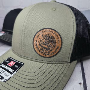 May include: A green and black trucker hat with a brown leather patch featuring the Mexican coat of arms. The patch has the text "Estados Unidos Mexicanos" around the edge. The hat has a black mesh back and a Richardson logo on the front.