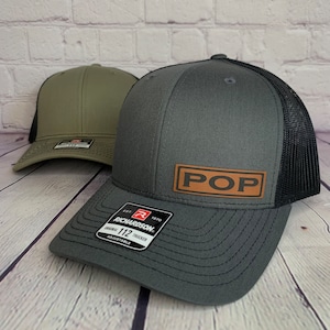 May include: A gray and black trucker hat with a brown leather patch that says "POP" on it. The hat is made by Richardson 112 and is adjustable.