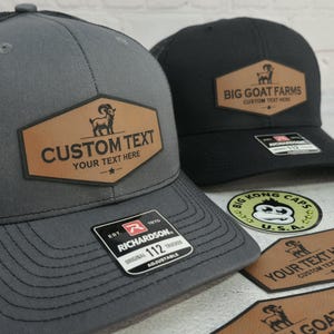 May include: Two trucker hats, one gray and one black, with custom leather patches. The gray hat has the text "CUSTOM TEXT YOUR TEXT HERE" and a goat graphic. The black hat says "BIG GOAT FARMS CUSTOM TEXT HERE". Both hats have a Richardson 112 tag.