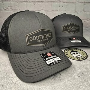 Puede incluir: Dos gorras de camionero grises con paneles de malla negra. Ambas gorras tienen un parche negro y plateado con el texto "Godfather Est. 2025". La gorra de la izquierda tiene una correa ajustable negra y roja con el texto "Richardson 112".