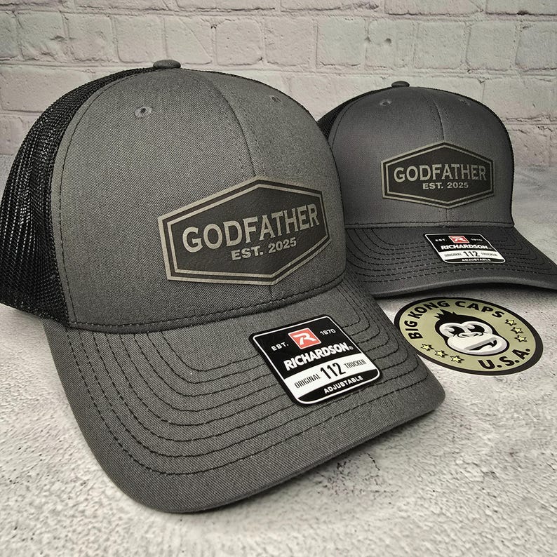 Puede incluir: Dos gorras de camionero grises con paneles de malla negra. Ambas gorras tienen un parche negro y plateado con el texto "Godfather Est. 2025". La gorra de la izquierda tiene una correa ajustable negra y roja con el texto "Richardson 112".