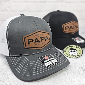 May include: Two trucker hats, one gray and one black, with a leather patch that reads "PAPA." The gray hat has a white mesh back and the text "MY FAVORITE PEOPLE Call Me PAPA Matthew & Megan." A "Big Kong Caps U.S.A." sticker is visible.