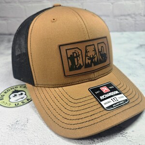 May include: Tan trucker hat with a black mesh back and a curved brim. The hat features a leather patch with the word "DAD" and hunting-themed silhouettes. The Richardson tag is visible on the brim.