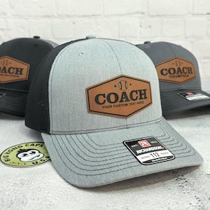 May include: Three baseball coach trucker hats. The hats are gray, black, and charcoal with a brown leatherette patch that says "COACH" and "YOUR CUSTOM TEXT HERE". The hats are adjustable and have a mesh back.