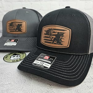 May include: Two Richardson 112 trucker hats. One is black with a gray mesh back, the other is gray with a black mesh back. Both have a brown leather patch with an archer and bow design. The hats are adjustable.