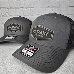 May include: Two gray trucker hats with black mesh back panels. The hats have a silver patch with the word "PAPAW" and the year established. The larger hat has "EST. 2025" and the smaller hat has "EST. 2022". The larger hat has a red and white label that says "RICHARDSON" and "112 TRUCKER".