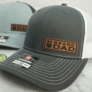 May include: Two trucker hats. The front hat is gray with a white mesh back and a brown leather patch that reads "BEARD DAD". The second hat is light gray with a black mesh back and a similar patch. Both hats have a curved brim and adjustable strap.