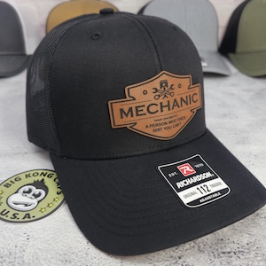 May include: Black trucker hat with a mesh back and a curved brim. The hat features a brown leather patch with the word "MECHANIC" and the definition: "A PERSON WHO FIXES SHIT YOU CAN'T". The hat is adjustable.