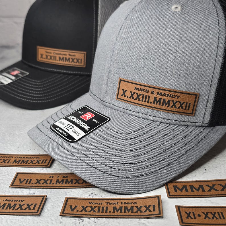 May include: A gray and black trucker hat with a brown leather patch that says "MIKE & MANDY X.XXIII.MMXXII". The hat is surrounded by several other brown leather patches with different dates and names.