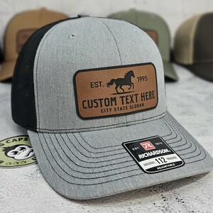 K&ouml;nnte beinhalten: Graue Trucker-Kappe mit schwarzem Mesh-R&uuml;cken und einem braunen Lederaufn&auml;her. Der Aufn&auml;her zeigt eine Pferdesilhouette, den Text "EST. 1995" und Platz f&uuml;r individuellen Text. Die Kappe hat einen gebogenen Schirm und einen verstellbaren Riemen.