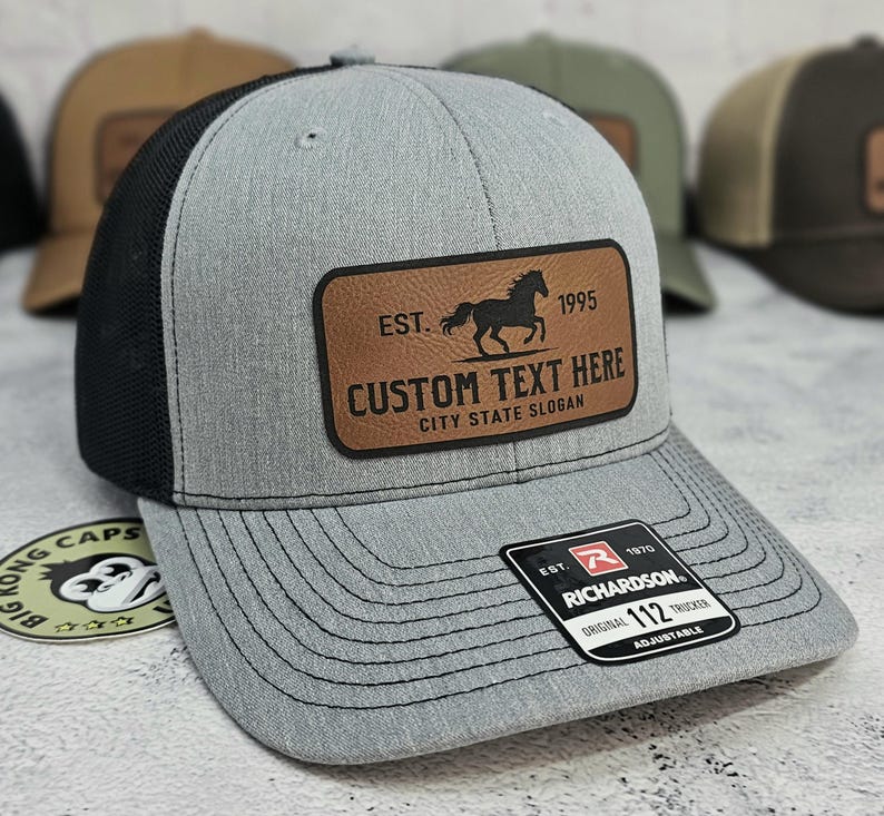 K&ouml;nnte beinhalten: Graue Trucker-Kappe mit schwarzem Mesh-R&uuml;cken und einem braunen Lederaufn&auml;her. Der Aufn&auml;her zeigt eine Pferdesilhouette, den Text "EST. 1995" und Platz f&uuml;r individuellen Text. Die Kappe hat einen gebogenen Schirm und einen verstellbaren Riemen.